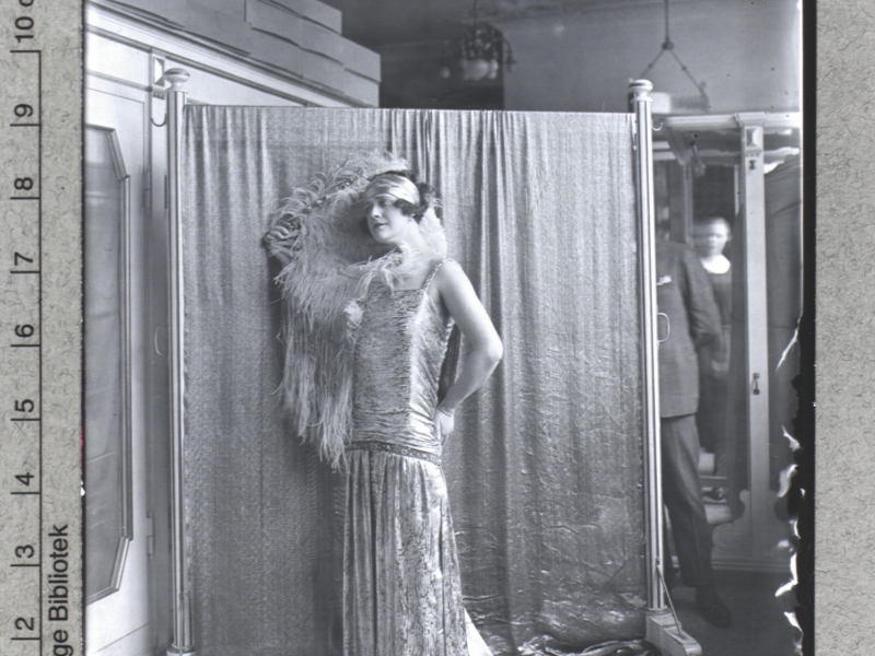 Kvinde poserer foran klæde iført lang kjole og fjer.