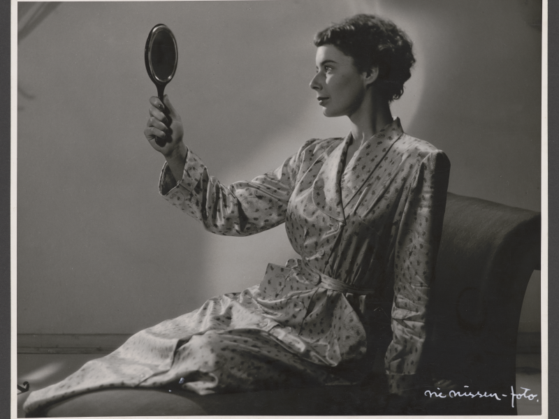 Kvinde ned og kigger sig selv i et lille håndholdt spejl. Hendes hår er sat op og bag hende oplyses en væg.