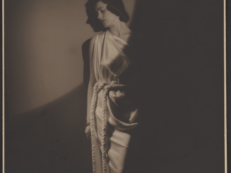 Kvinde står op ad en væg. Hun er klædt i en græsk-inspireret kjole med reb som bælte. Et enkelt lys skinner på hendes ansigt.