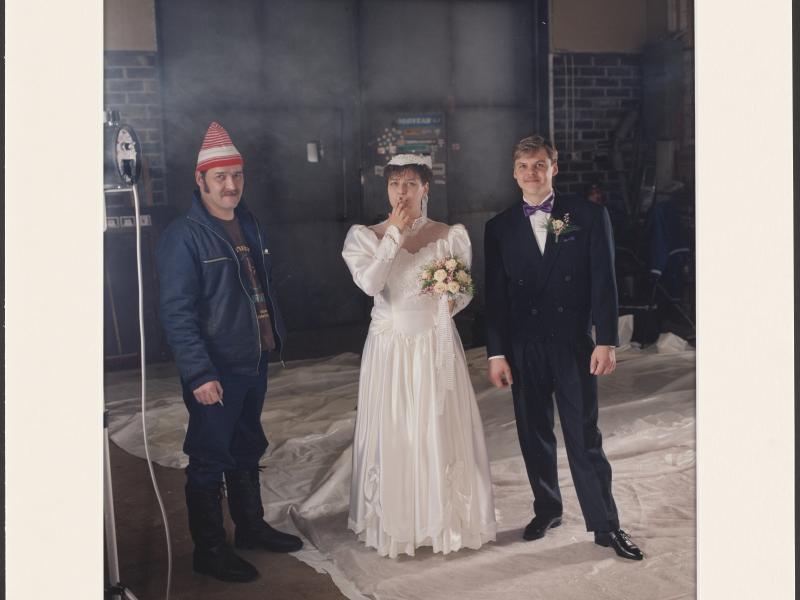 Et brudepar står ved siden af en mand i denimjakke og rød hat