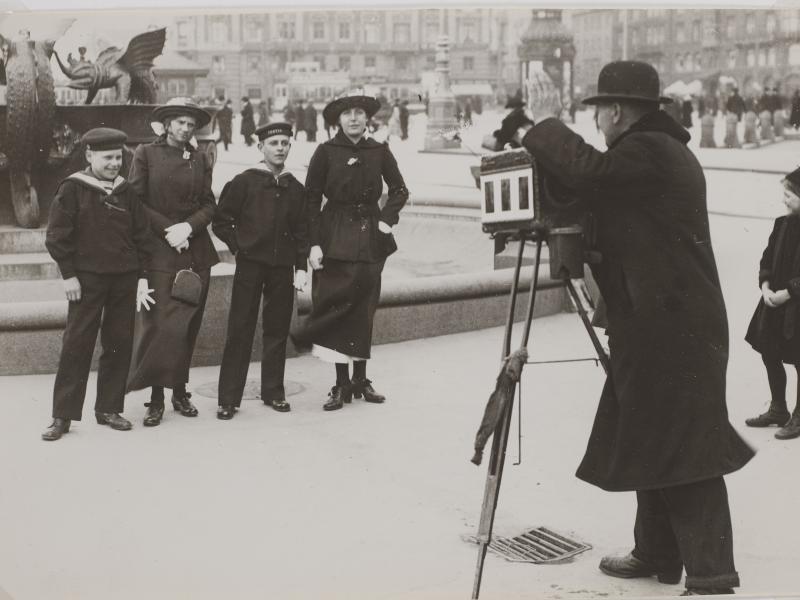 Fotograf med gammeldags kamera tager billede af fire mennesker foran et springvand. To kvinder og to mænd.