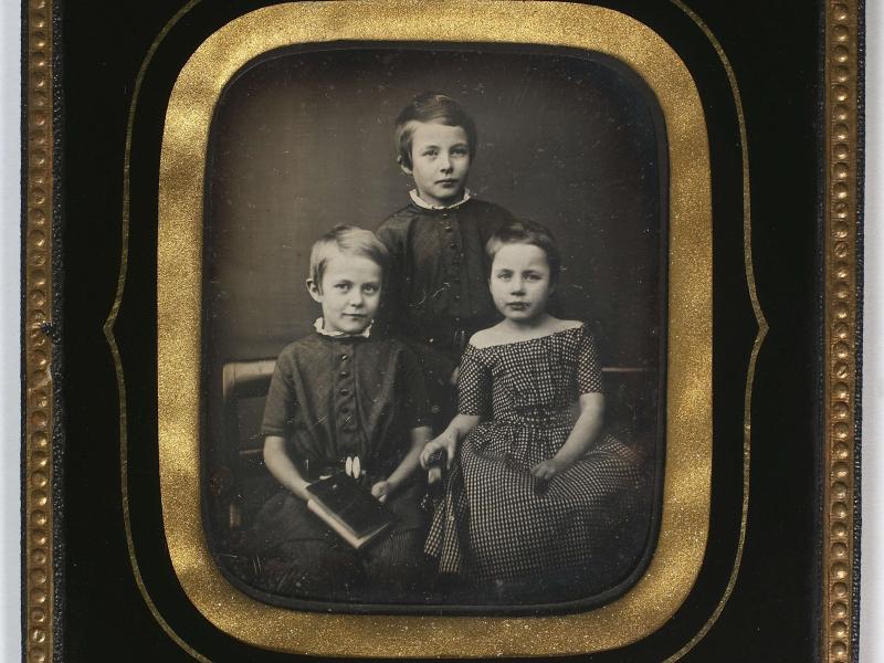 Mørkt billede af tre børn i pænt tøj. Billedet er i en flot ramme.