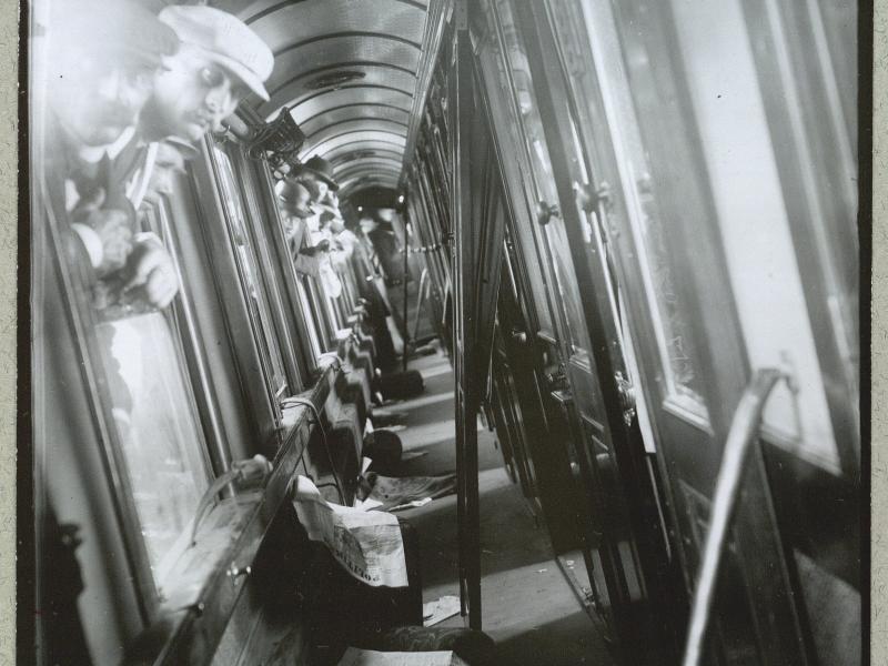 Flere mænd stikker hovedet ud af vinduer til gangen på et tog. På jorden ligger der aviser.