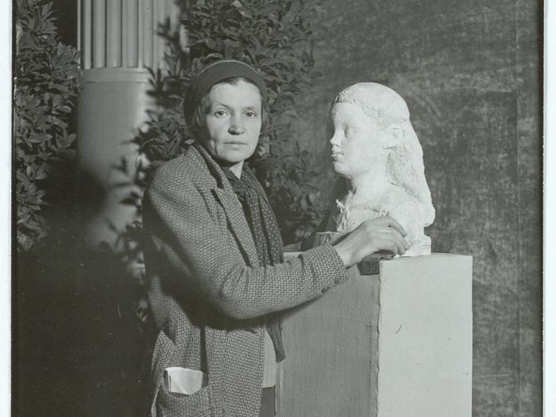 Billedhuggeren Astrid Noack står ved siden af en statur af en kvindes ansigt. Noack bærer en frakke og hue.