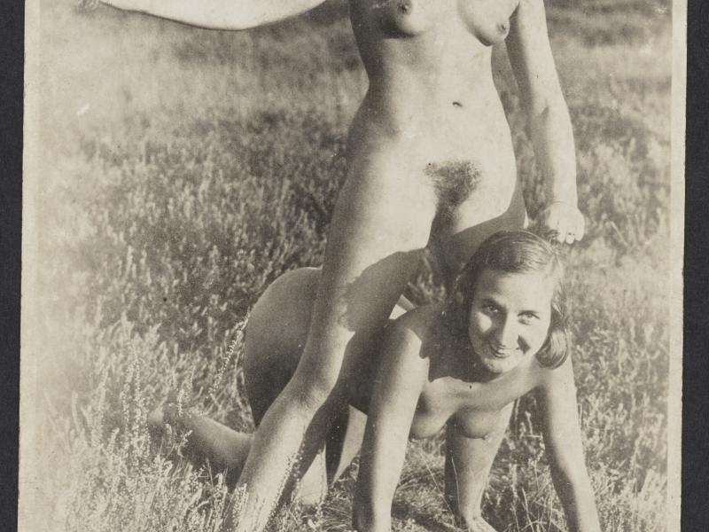 En nøgen kvinde på alle fire på en eng, mens en anden nøgen kvinde står over hende med en buket i hånden