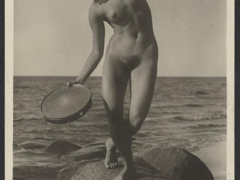 Nøgen kvinde på en sten ved havet med en tamburin i hånden