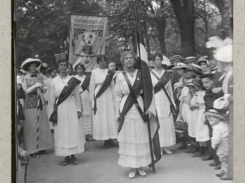 Kvindetoget står med banner og dannebrog. De er klædt i hvide kjoler.