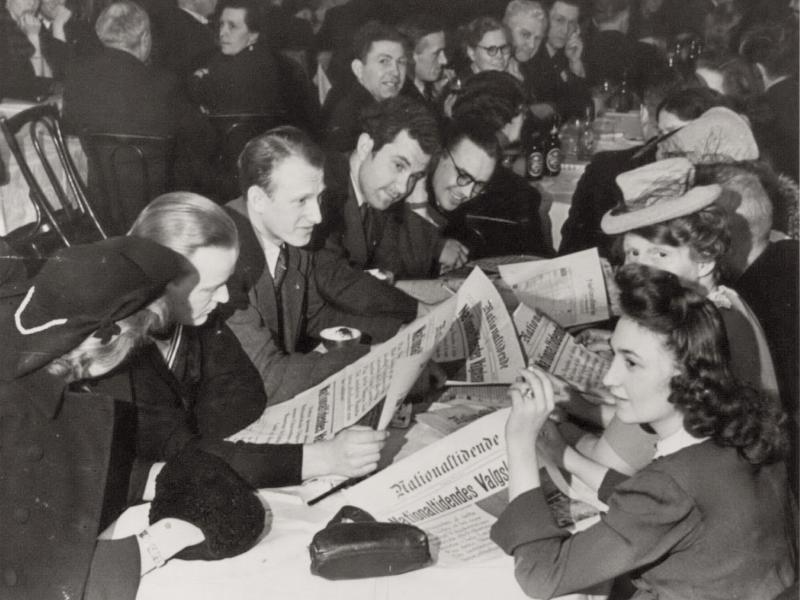 Mænd og kvinder sidder ved langbord med avisen National Tidende