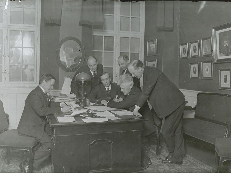 Mænd står og sidder ved skrivebord med masser af papirer.