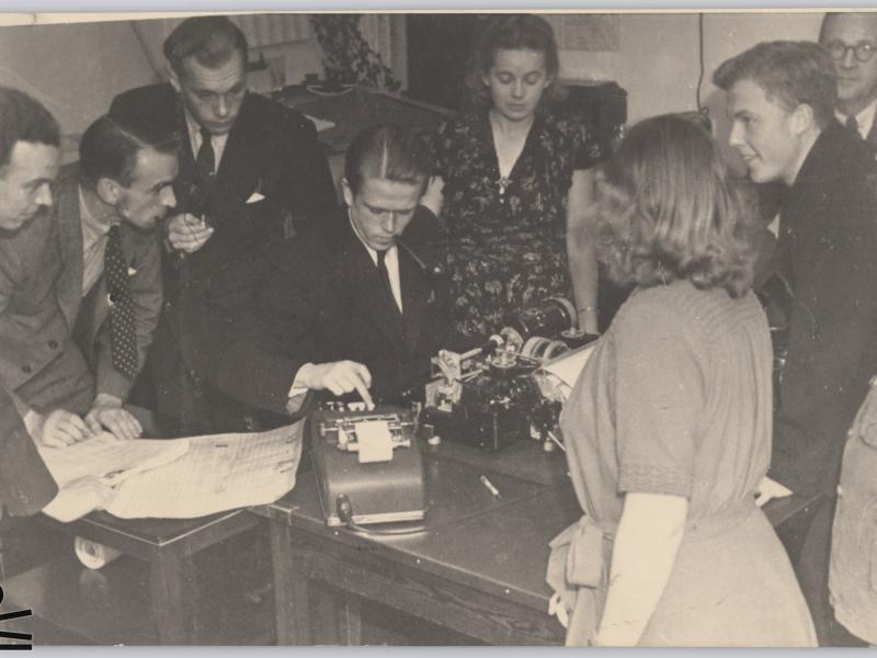 Man is bent over typewriter. Women and men watch.