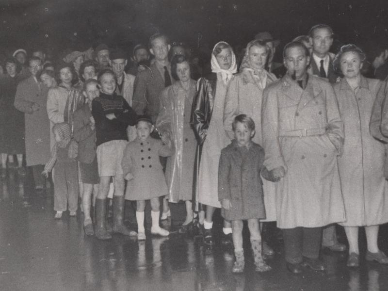 Women and men in trench coats stand in a long line. Some have children with them.