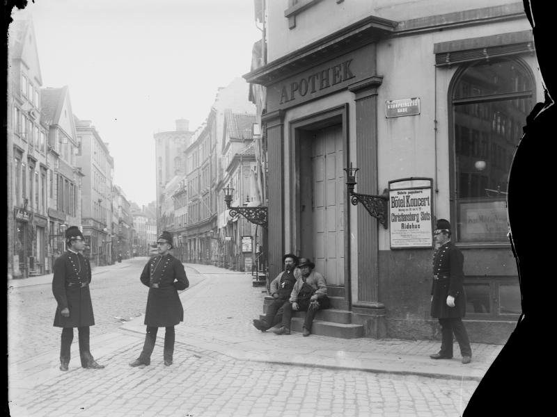 Tre politimænd står på et gadehjørne. To mænd sidder på trappen ved et apotek