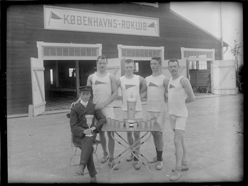 Fire mænd klædt i hvidt og en mand klædt i sort foran Københavns Roklub med pokaler