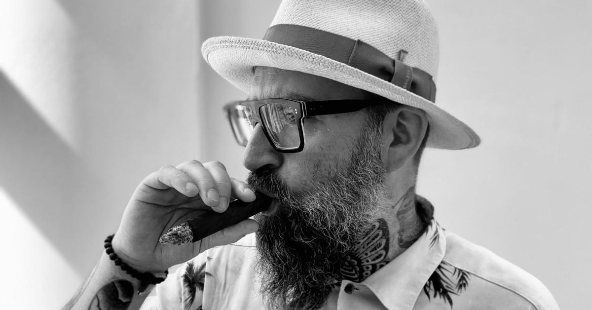 Black and white picture of the poet Claus Høxbroe in summer shirt and hat, smoking a cigar.