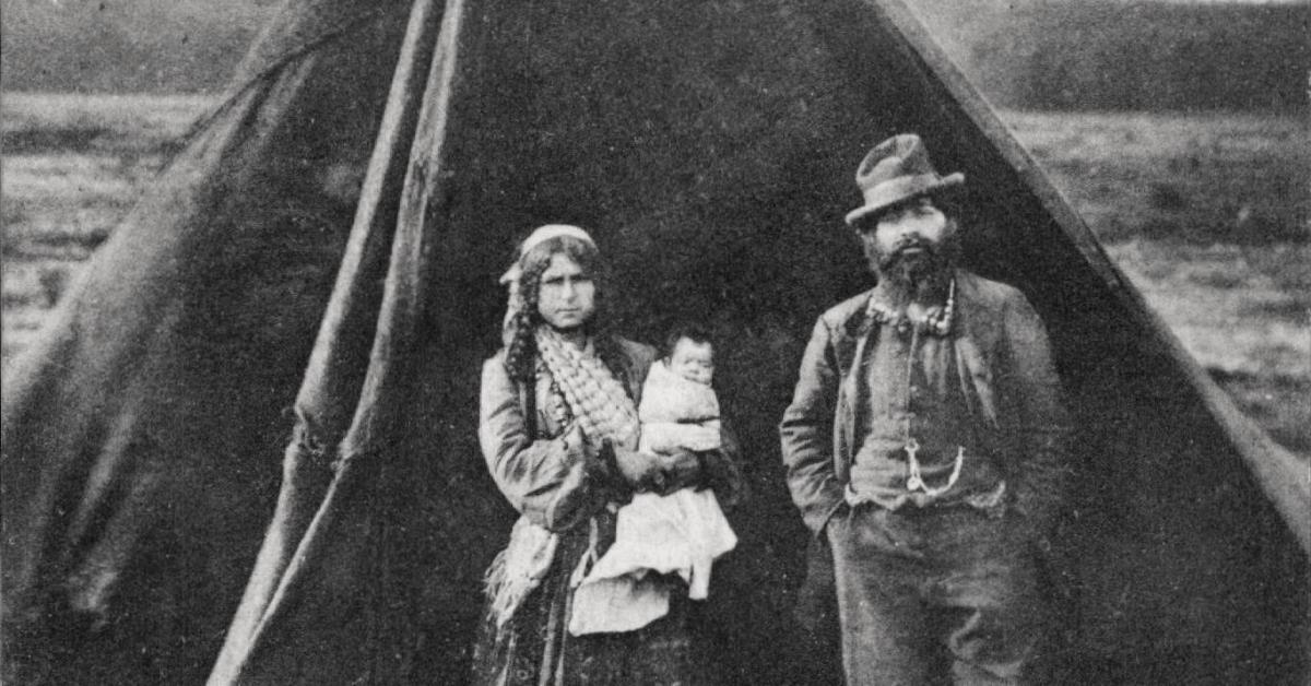 En familie bestående af en mor, en far og en baby fra en såkaldt ”Zigøjnerlejr”. Moren holder det lille barn, der er svøbt ind. De står foran et telt iført mange lag tøj og ser ind i kameraet.